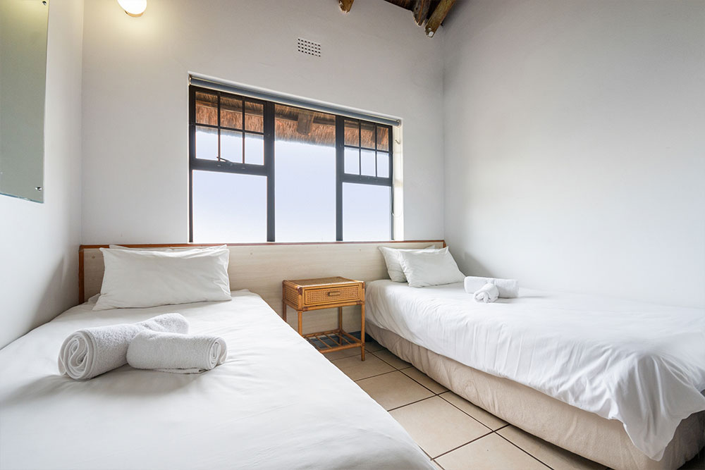 An upgraded one-bedroom features two single beds with crisp white linens and rolled towels. A small wooden nightstand rests between the beds, positioned under a window that fills the space with natural light. @De Bakke Santos Mossel Bay