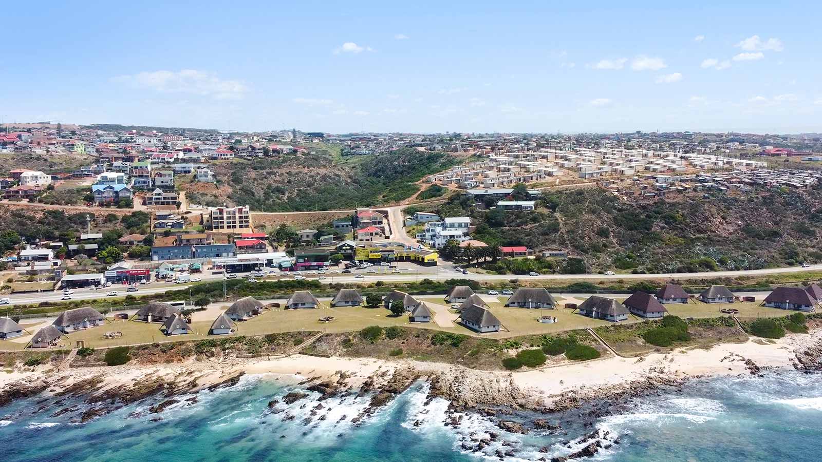 A coastal landscape with thatched-roof huts along the shoreline, a sandy beach, clear turquoise water, and a residential town with colorful houses and buildings on hilly terrain—often praised in customer reviews for its stunning beauty. @De Bakke Santos Mossel Bay