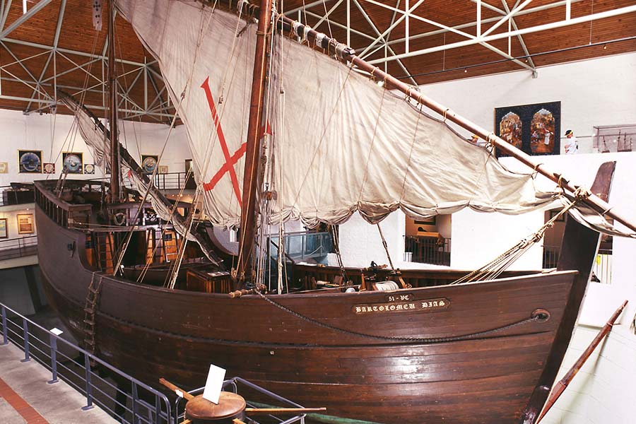 A large wooden replica of a historic sailing ship with a single sail marked by a red cross, displayed indoors at a museum with railings and art on the walls. @De Bakke Santos Mossel Bay