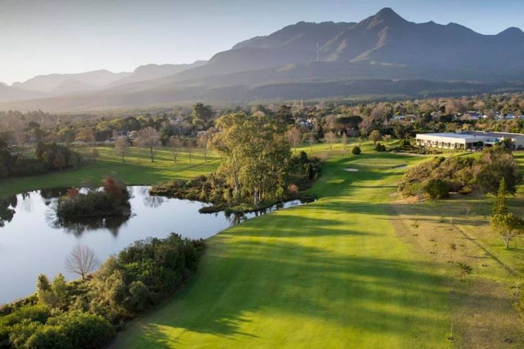 A scenic golf course with green fairways, a pond, and scattered trees, set against a backdrop of mountains under a clear sky. A building is visible on the right side near the edge of the course. @De Bakke Santos Mossel Bay