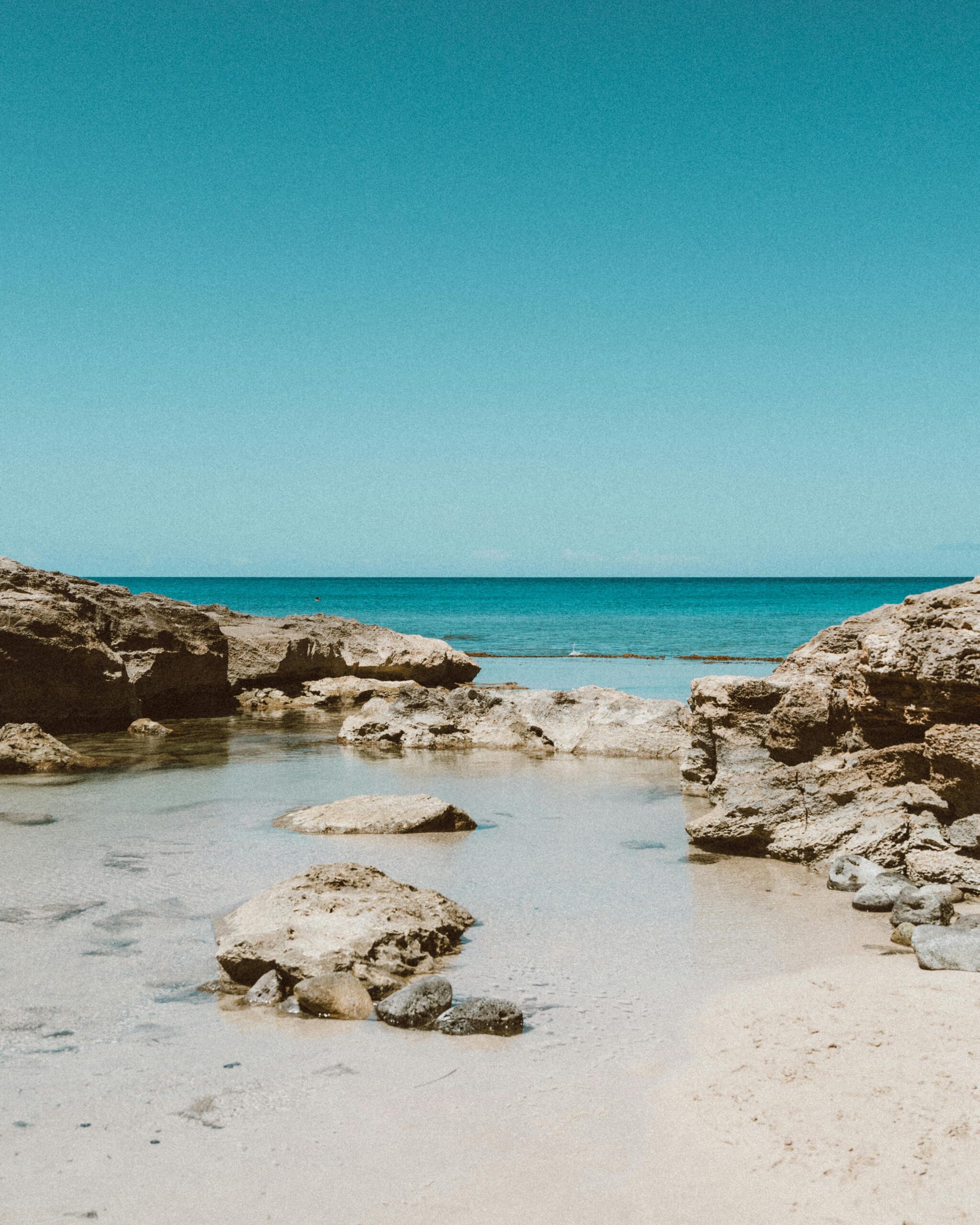 Rocky shoreline with clear, shallow water and scattered rocks, opening up to a calm blue sea under a clear sky. The scene is peaceful and sunlit, with no people visible. @De Bakke Santos Mossel Bay