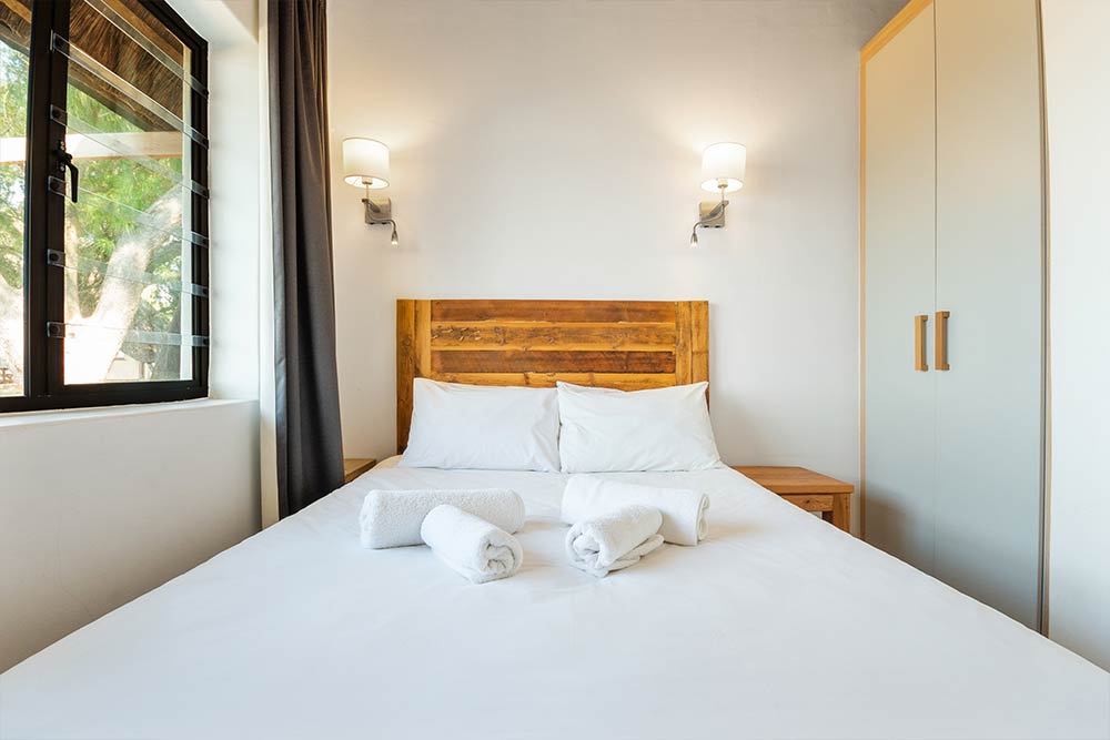 A neatly made bed with white linens and three rolled white towels on top, positioned between wooden nightstands, under two wall-mounted lamps, next to a window and a wardrobe in a bright, modern room. @De Bakke Santos Mossel Bay