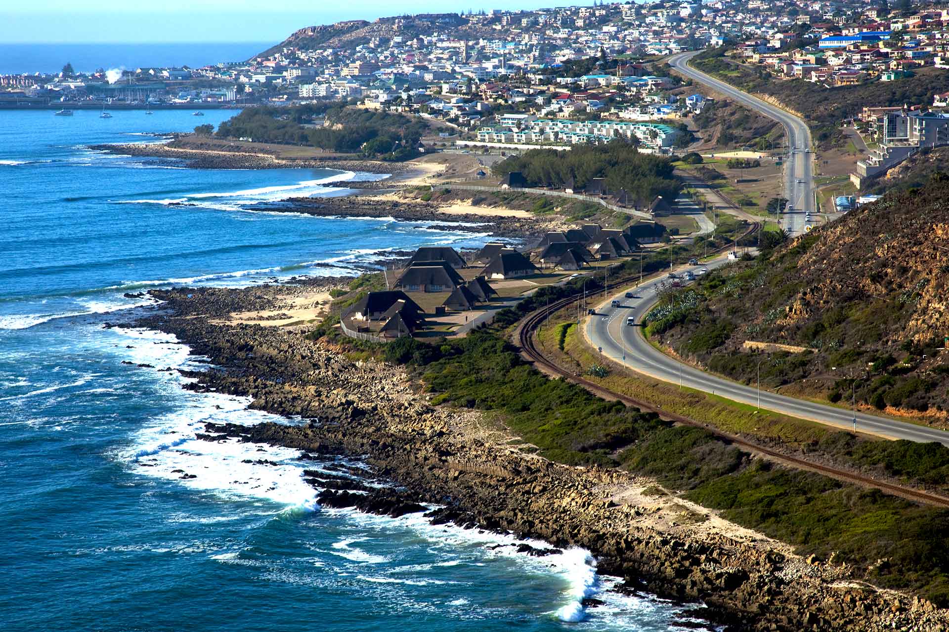 A coastal town with houses and roads along a rocky shoreline, waves crashing against the rocks, and hills in the background under a clear blue sky—perfect for self catering and camping accommodation Mossel Bay. @De Bakke Santos Mossel Bay
