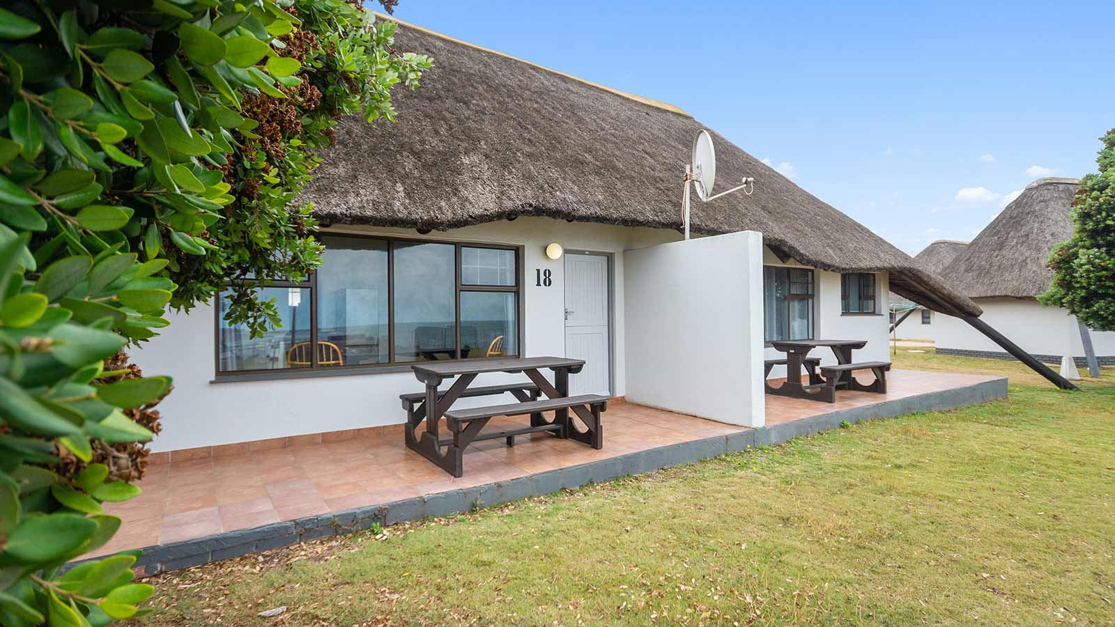 Single-story Beach Chalet with white walls and a thatched roof, featuring two outdoor picnic tables on a tiled patio. The cottage is surrounded by grass, with another similar building visible in the background. @De Bakke Santos Mossel Bay