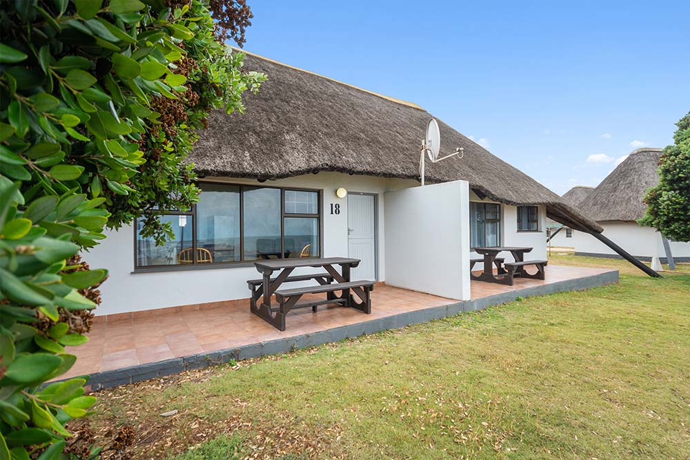 A thatched-roof white Two Bedroom Chalet, labeled “18,” features two wooden picnic tables on a tiled patio. The chalet is surrounded by green grass and bushes under a clear blue sky. @De Bakke Santos Mossel Bay