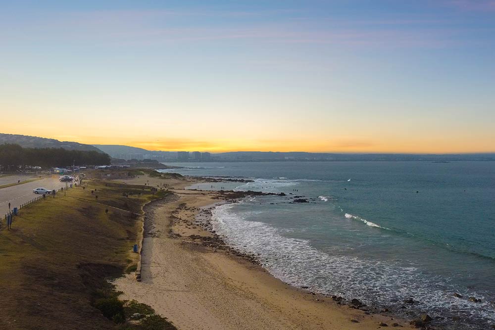 A peaceful beach scene at sunset with gentle waves, sandy shore, scattered rocks, and a road running parallel to the coastline. A few people walk as soft orange and blue hues fill the sky—perfect for a gallery slider showcasing tranquil moments. @De Bakke Santos Mossel Bay