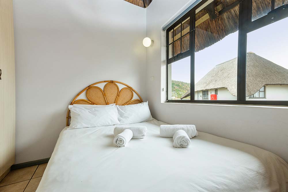 A neatly made bed with white linens and three rolled towels sits by a window with a thatched roof view outside. This upgraded three bedroom chalet features light walls, a rattan headboard, and soft natural light. Sleeps 5. @De Bakke Santos Mossel Bay