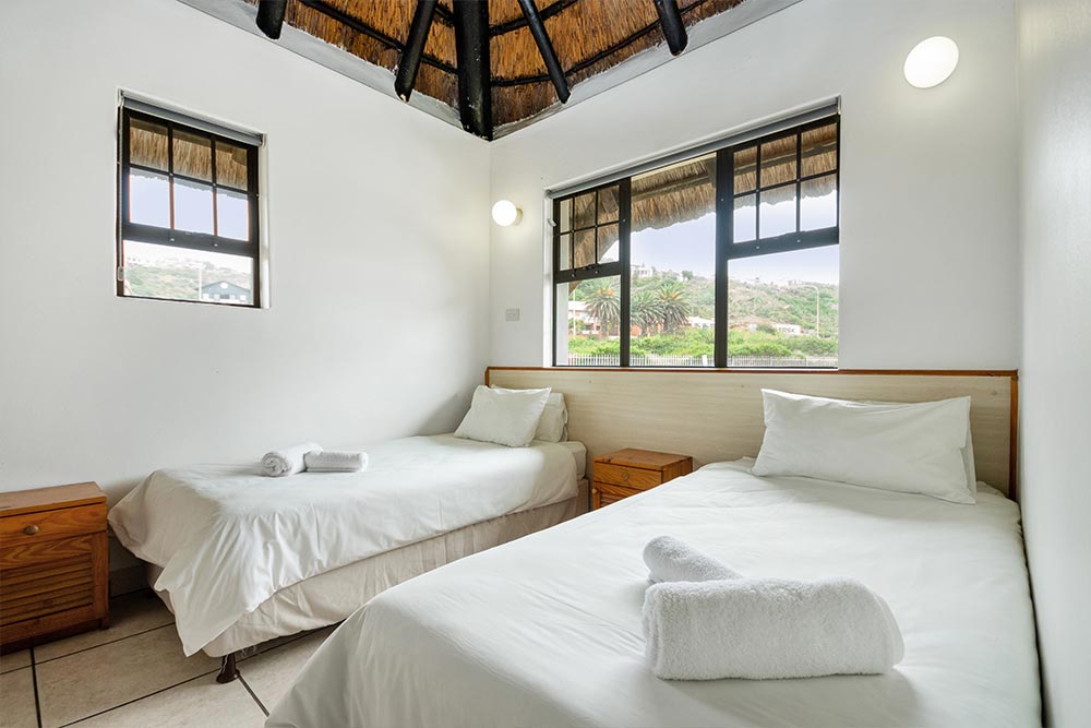 A bright Upgraded Chalet room with two single beds, white bedding, rolled towels, wooden nightstands, and large windows showing a scenic outdoor view. The ceiling features exposed wooden beams. @De Bakke Santos Mossel Bay