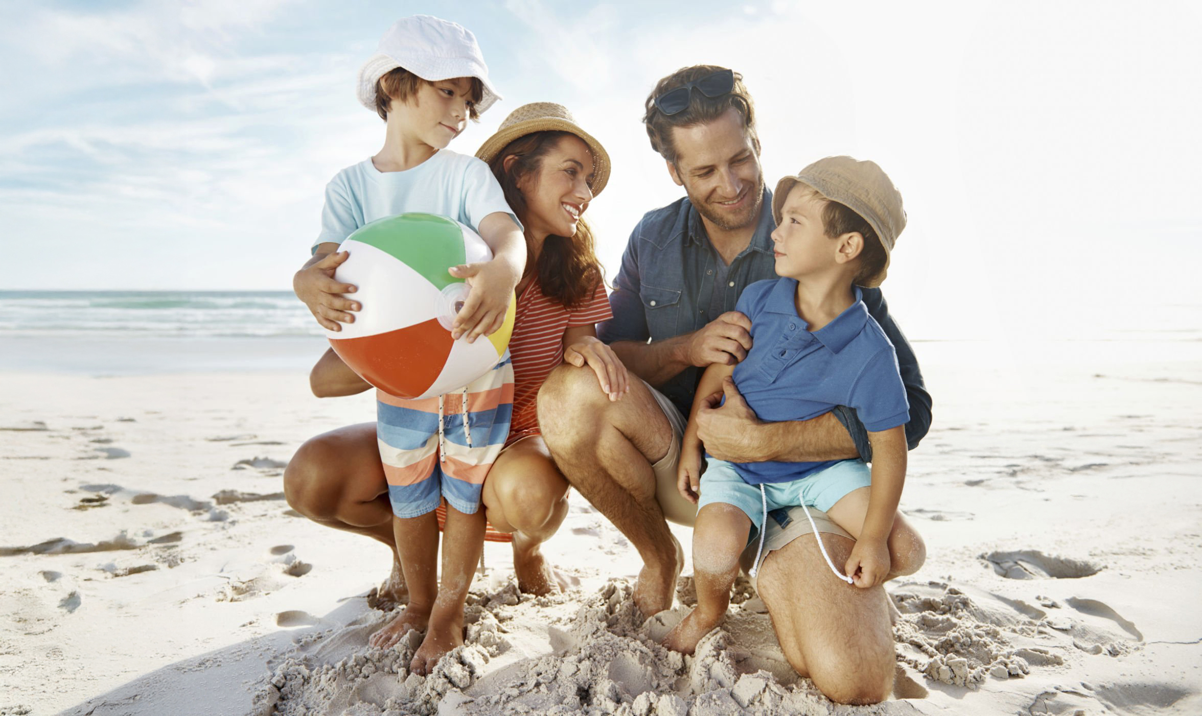 A smiling family of four—two adults and two young children—stands together on a sandy beach. One child holds a colorful beach ball. The ocean and blue sky are in the background, as they enjoy their vacation specials, looking happy and relaxed. @De Bakke Santos Mossel Bay