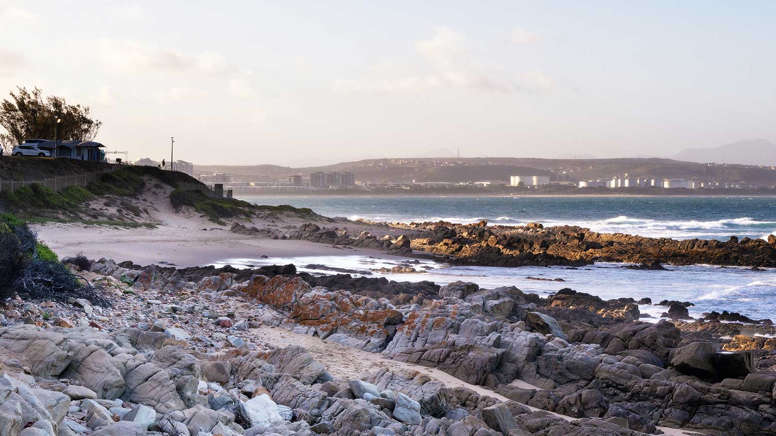 Rocky shoreline with scattered vegetation leading to a sandy beach. Waves crash against the rocks, and buildings are visible across the bay under a partly cloudy sky. A few vehicles are parked near the edge of the beach in these De Bakke Santos images. @De Bakke Santos Mossel Bay
