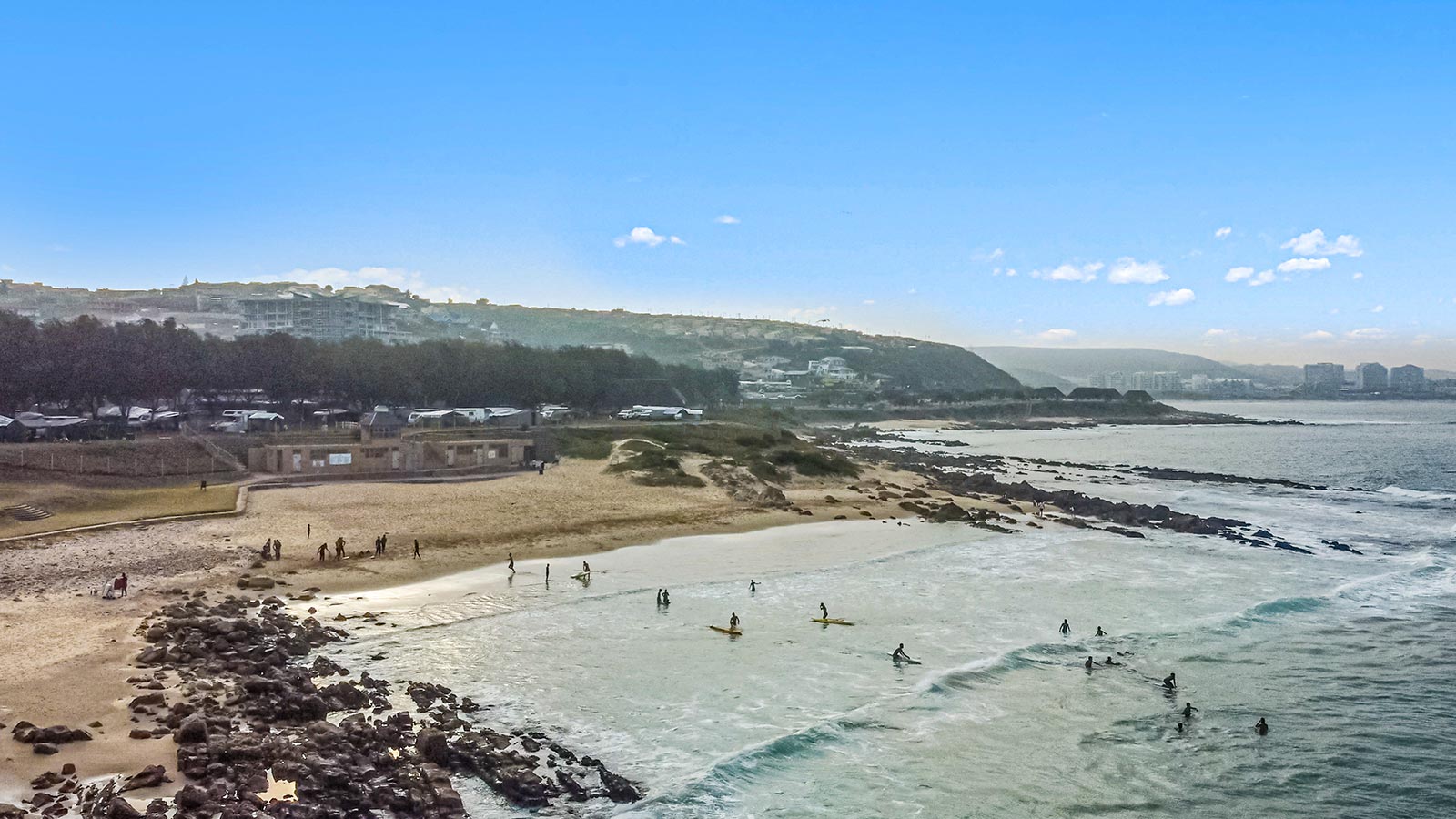 A beach scene with several people swimming and surfing in the ocean, while others walk or relax on the sandy shore; rocky areas and buildings are visible in the background under a clear blue sky, making it perfect for enjoying summer specials. @De Bakke Santos Mossel Bay