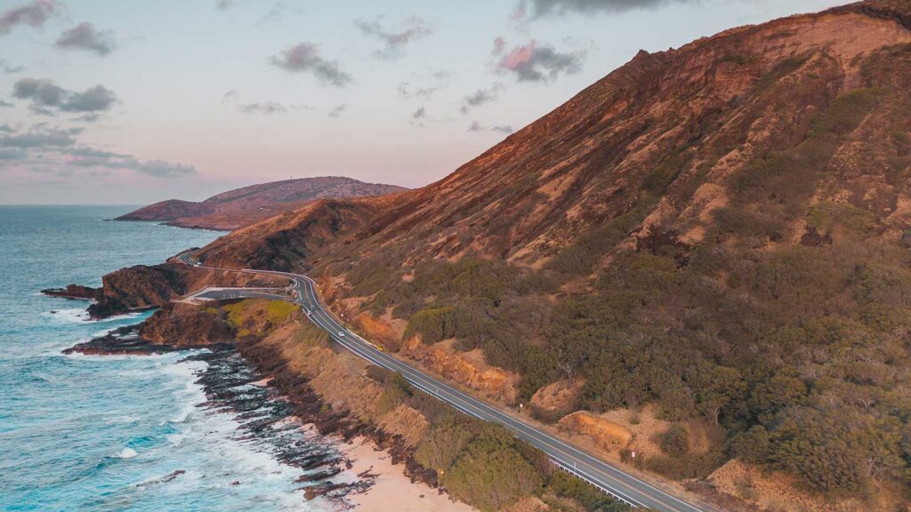 A winding coastal road runs alongside steep, rocky hills and the ocean, with waves crashing near self catering beach front chalet's in Mossel Bay under a partly cloudy sky at sunset. @De Bakke Santos Mossel Bay