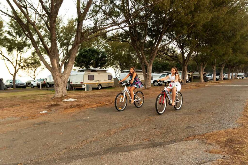 Two girls ride bicycles on a paved road through a campground at De Bakke Santos, with caravans and cars parked under tall trees. The relaxed, outdoorsy scene captures the essence of De Bakke Santos images. @De Bakke Santos Mossel Bay