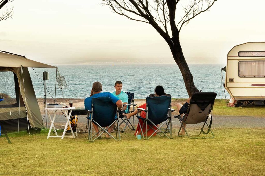 Four people sit in camping chairs near tents and a caravan, with trees and the ocean in the background, enjoying a relaxed outdoor setting—capturing the essence of De Bakke Santos images. @De Bakke Santos Mossel Bay