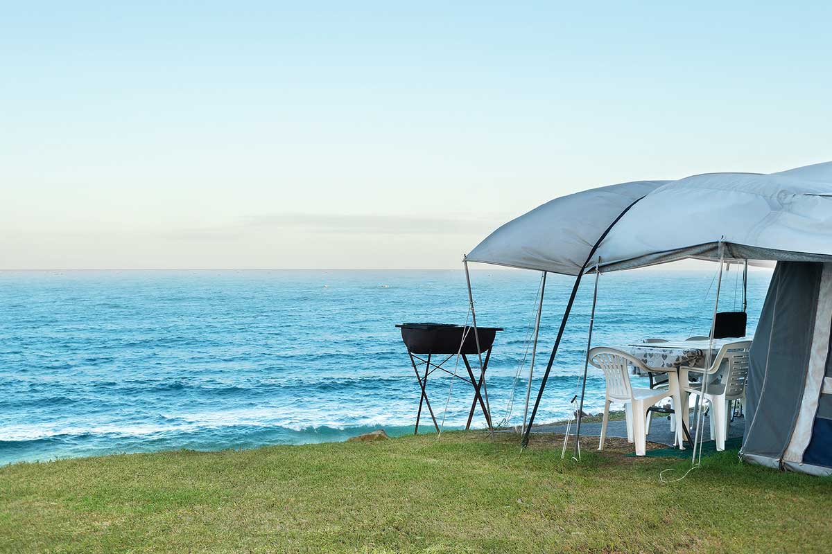 A tent with a canopy, white plastic chairs, and a table is set up on grassy ground by the ocean. A barbecue grill stands nearby with clear blue water and sky in the background—classic De Bakke Santos Images showcasing resort photos. @De Bakke Santos Mossel Bay