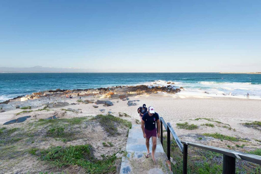 A person in shorts and a cap walks up stone steps from a sandy beach with scattered rocks at De Bakke Santos, grassy dunes nearby, and waves crashing under a clear blue sky—perfect for resort photos. @De Bakke Santos Mossel Bay
