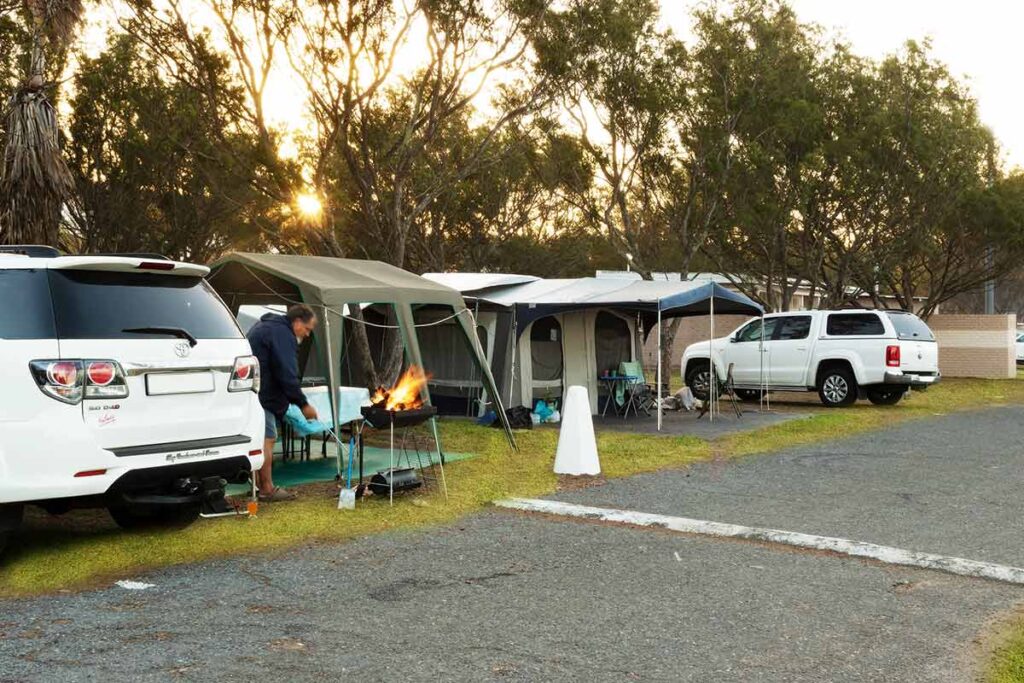 Two white SUVs are parked beside tents in a campsite, as seen in De Bakke Santos Images. A person tends to a campfire on a grill, surrounded by trees, folding chairs, and camping gear, with the sun setting in the background. @De Bakke Santos Mossel Bay