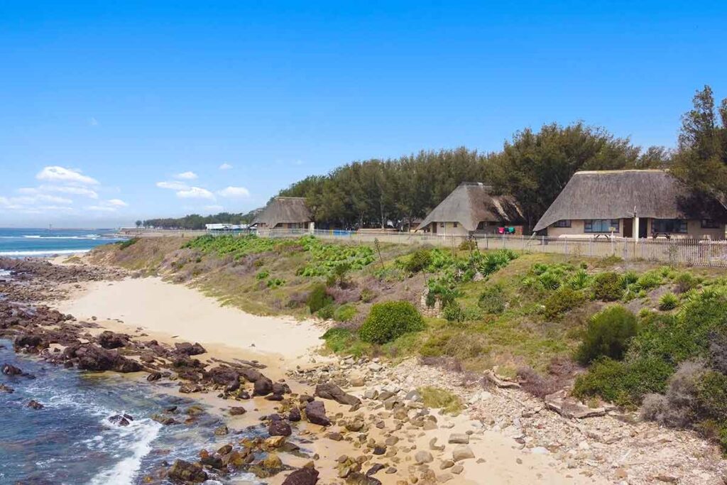 Thatched-roof cottages line a sandy and rocky coastline, bordered by green shrubs and trees, with blue ocean waves and a clear sky in the background—capturing the charm seen in De Bakke Santos Images. @De Bakke Santos Mossel Bay
