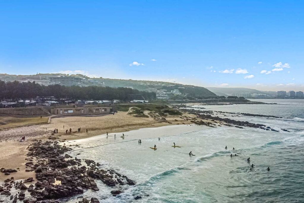 A beach with light waves and scattered surfers in the water, people walking along the sandy shore, rocks in the foreground, and a coastal town with green hills under a clear blue sky—perfect for De Bakke Santos Images. @De Bakke Santos Mossel Bay