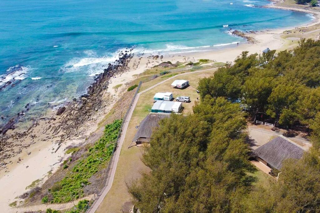 Aerial view of De Bakke Santos Images shows a coastal area with sandy beach, rocky shoreline, blue ocean waves, and several small buildings with thatched roofs surrounded by trees and grassy land. A path runs parallel to the shoreline. @De Bakke Santos Mossel Bay