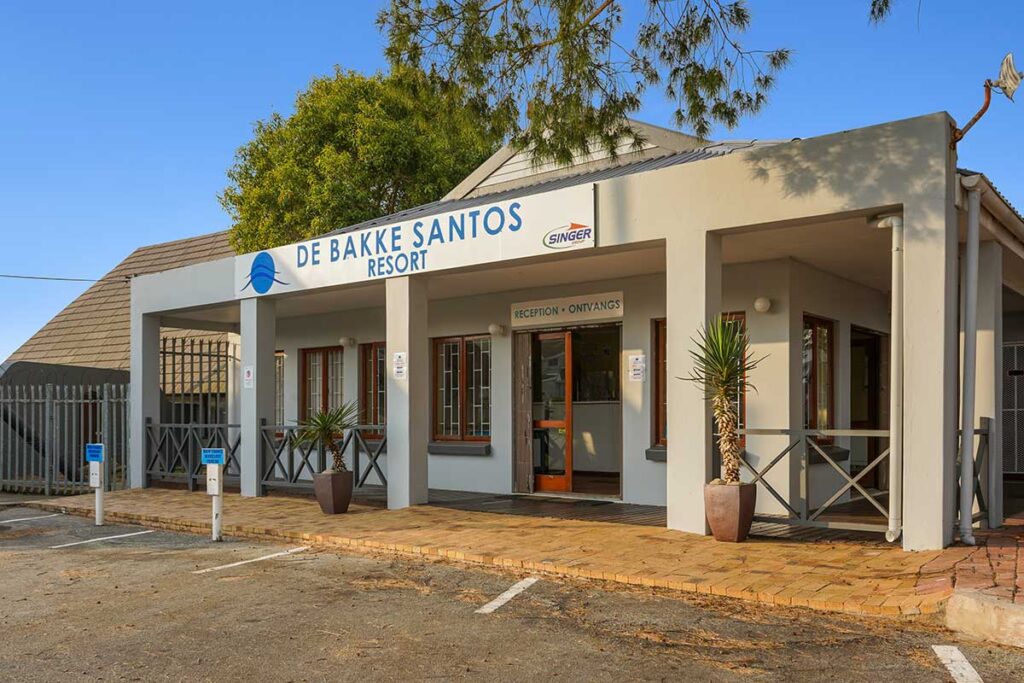 Single-story building with a sign reading "De Bakke Santos Resort" above the entrance. The front features a covered porch, potted plants, and parking spaces—see more in our De Bakke Santos Images showcasing trees and sunlight in the background. @De Bakke Santos Mossel Bay