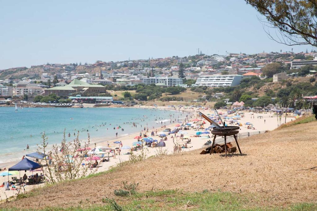 A busy beach with many people, colorful umbrellas, and calm blue water. In the foreground, an empty chair and a small grill sit on a grassy slope—reminiscent of De Bakke Santos images—with a coastal town in the background. @De Bakke Santos Mossel Bay