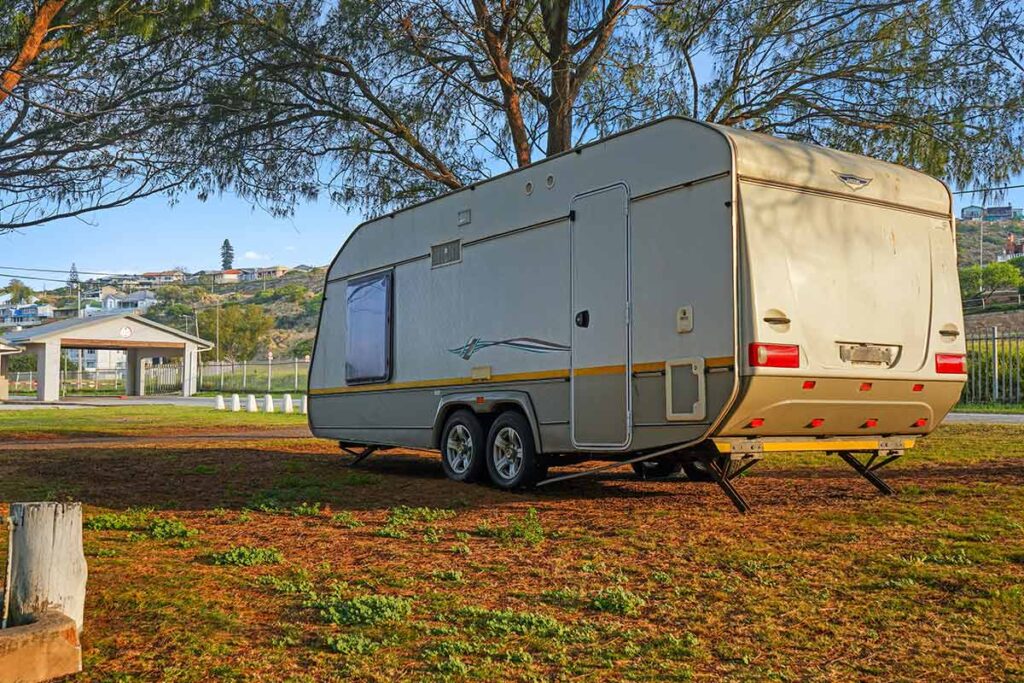 A beige travel trailer is parked on a grassy area under trees in a campground, with sunlight casting shadows and a small building visible in the background, capturing the tranquil vibe found in De Bakke Santos Images. @De Bakke Santos Mossel Bay