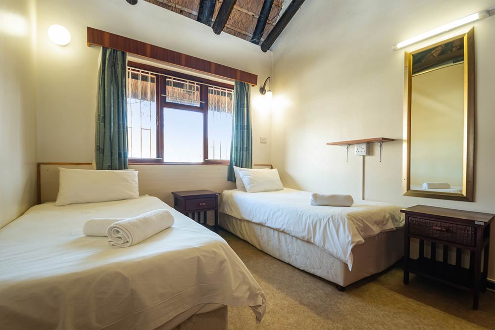A simple, clean Two Bedroom Chalet room with two single beds, each with white bedding and a rolled towel. There are two bedside tables, a window with green curtains, and a wall mirror with a shelf beneath it. Exposed wooden beams on the ceiling add charm. @De Bakke Santos Mossel Bay