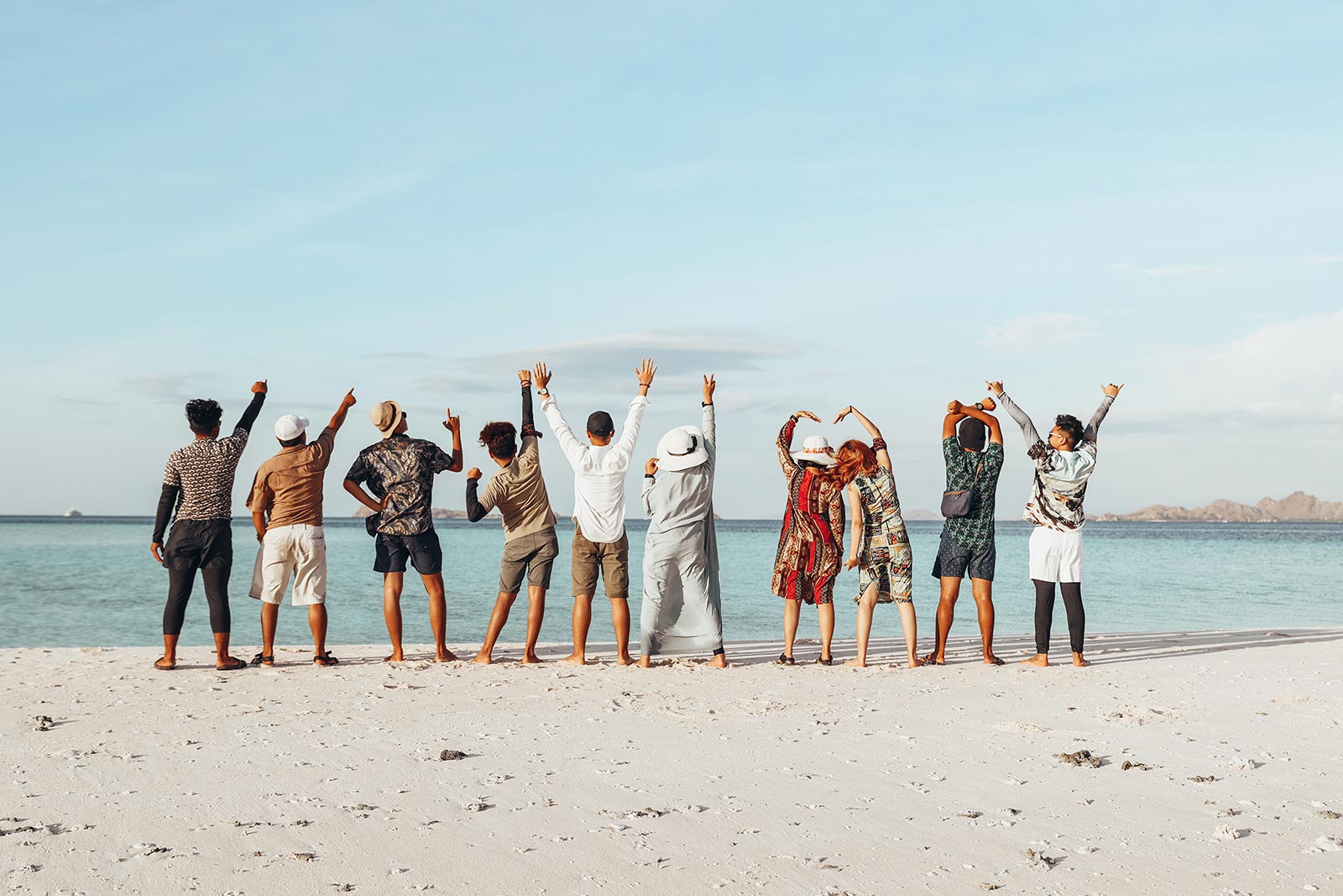Ten people stand in a row on a sandy beach along the Garden Route, arms raised and backs to the camera, enjoying a bright group getaway on a sunny day with clear blue skies. @De Bakke Santos Mossel Bay