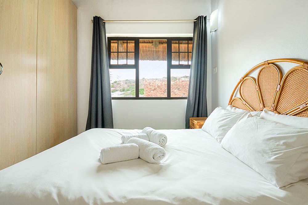 An upgraded, bright, minimal bedroom in the Three Bedroom Chalet featuring a large bed with white linens, rattan headboard, wooden nightstand, two rolled towels, and a window with dark curtains overlooking tiled rooftops and greenery. Sleeps 6. @De Bakke Santos Mossel Bay