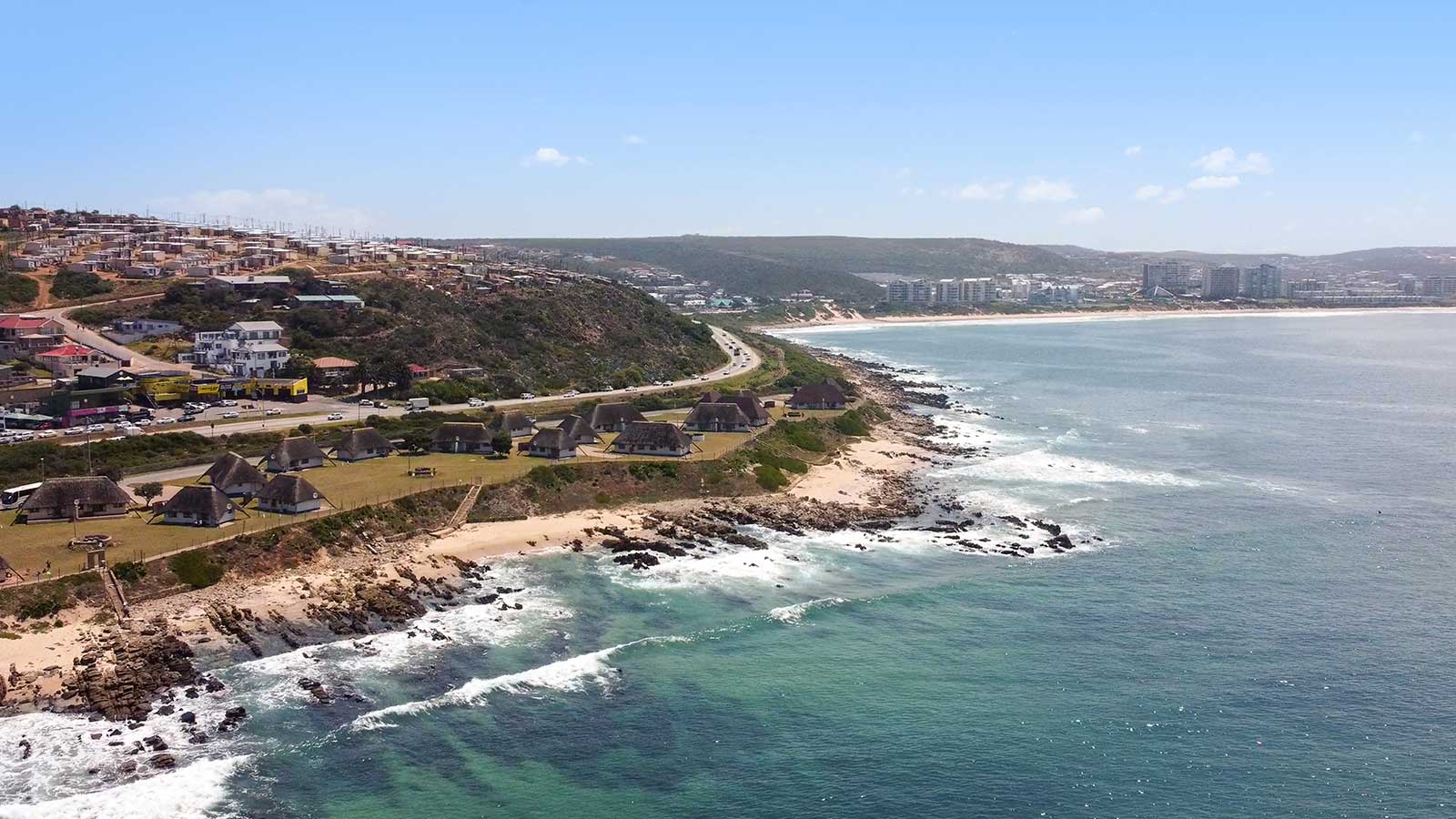 Aerial view of a coastal town with houses on a hillside, a road along the shoreline, rocky beaches, and waves crashing against the shore—plus nearby attractions and city buildings visible in the distance under a clear blue sky. @De Bakke Santos Mossel Bay