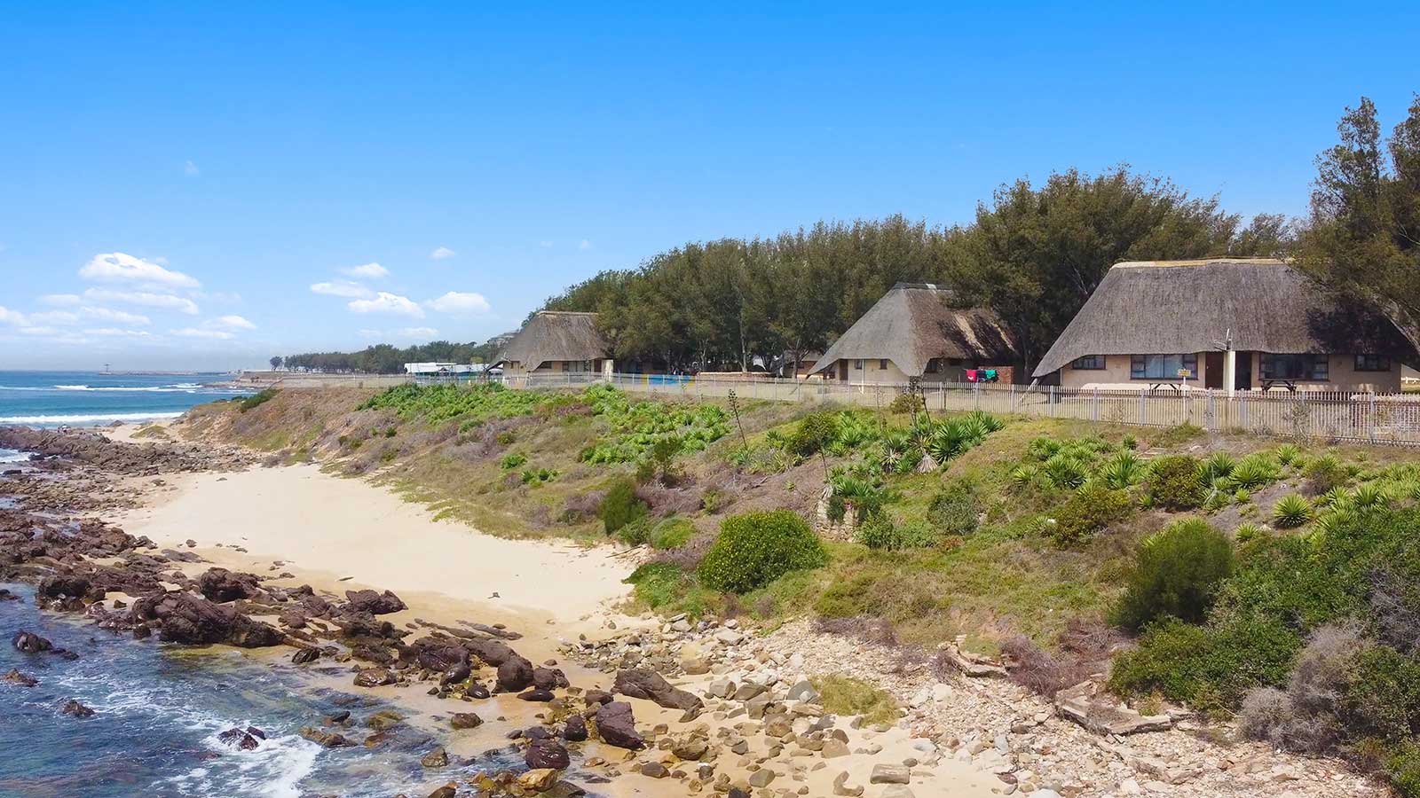 A coastal scene with thatched-roof cottages near the shoreline, surrounded by greenery and a rocky beach, offers stunning ocean views under a clear sky—perfect for a getaway with seasonal rates available. @De Bakke Santos Mossel Bay