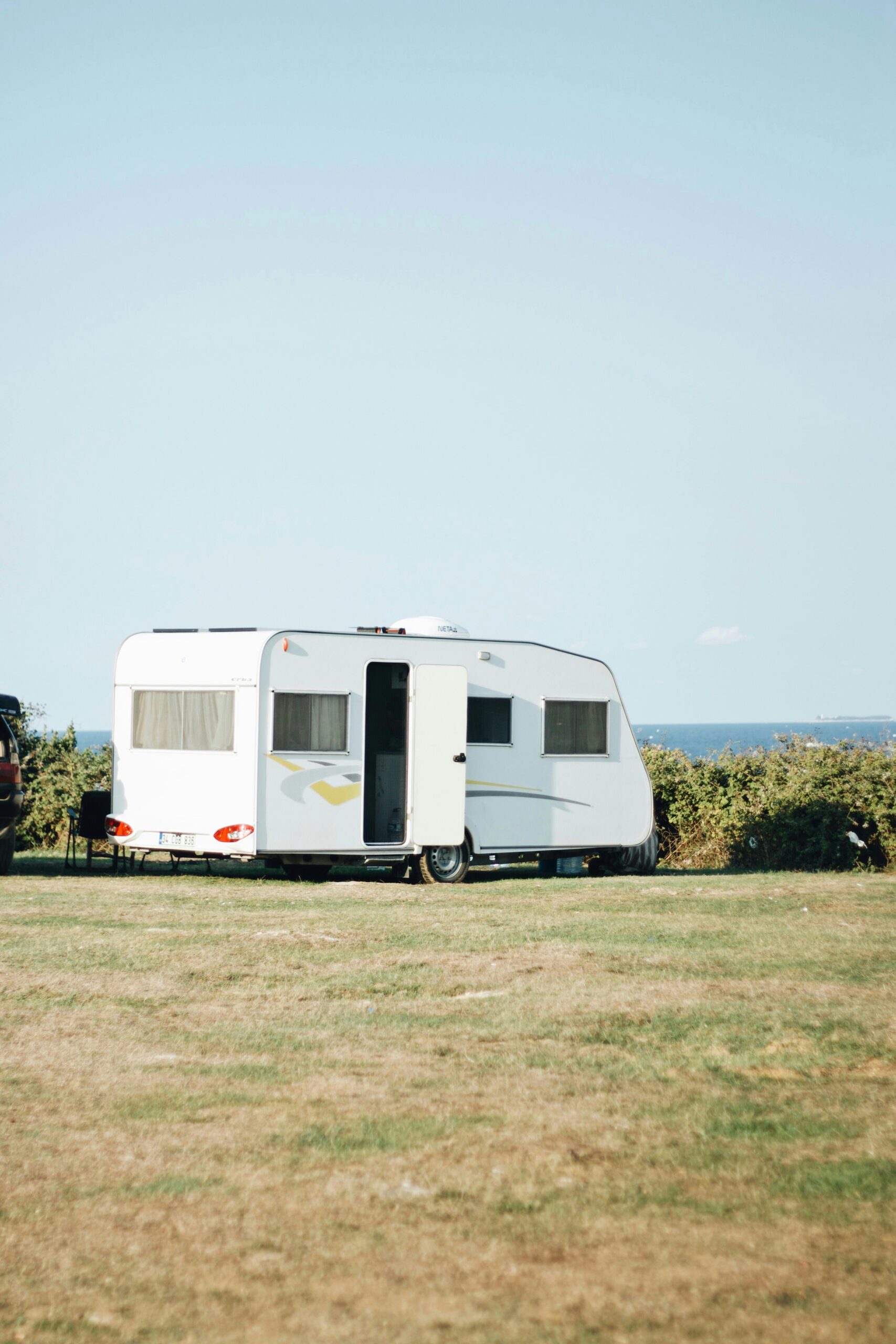 A white caravan with an open door is parked on a grassy field near green bushes. The sky is clear and blue, the sea visible in the background—perfect for beachfront caravan hire Mossel Bay offers. @De Bakke Santos Mossel Bay