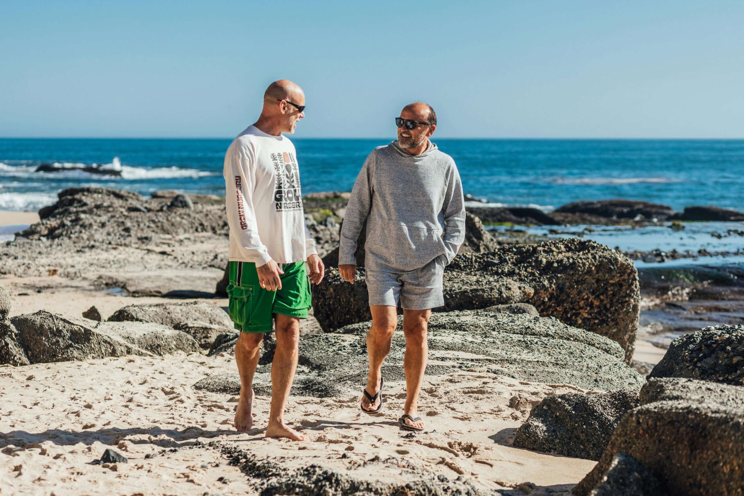 Two men in casual clothing walk barefoot on a sandy beach near large rocks, smiling and talking about the Pensioner Special, with blue ocean waves and a clear sky in the background. @De Bakke Santos Mossel Bay