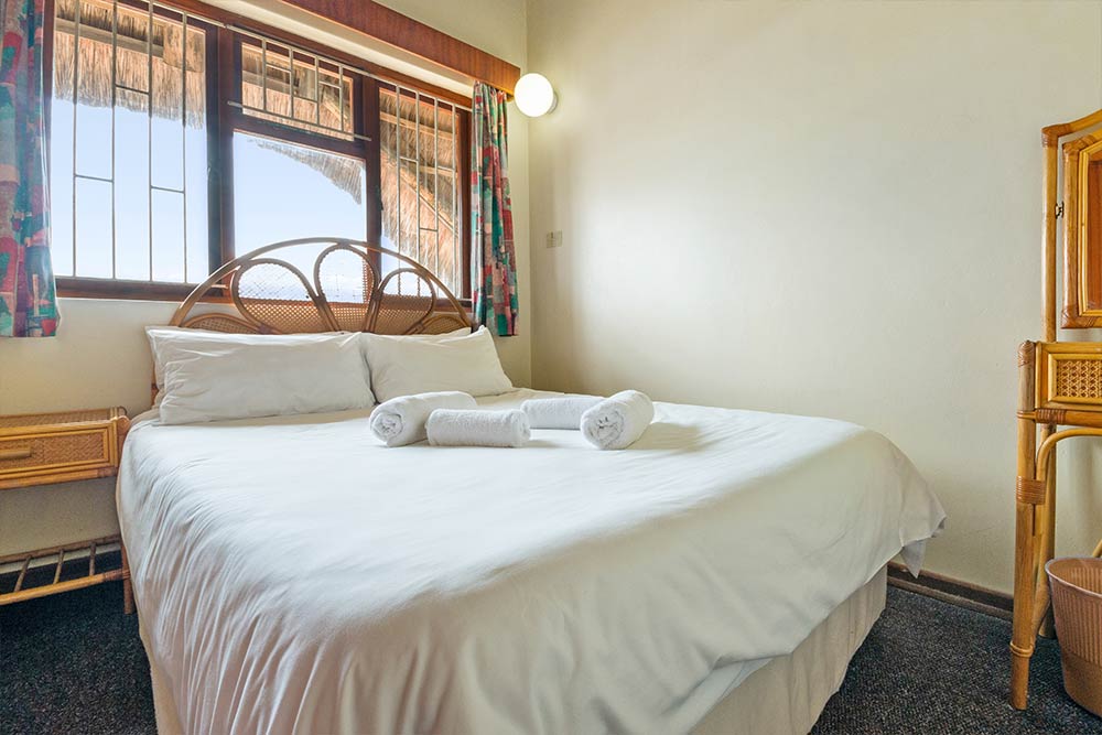 A neatly made bed with white linens and three rolled towels on top, placed in a bright Three Bedroom Chalet with a wooden headboard, a window with bars and curtains, and light-colored furniture. Sleeps 6 comfortably. @De Bakke Santos Mossel Bay