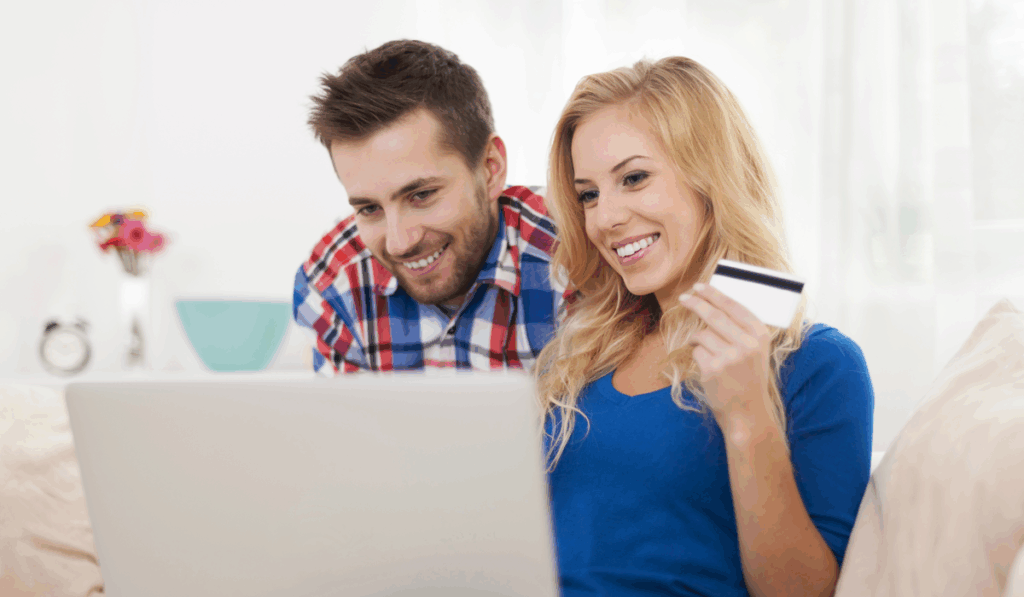 A smiling couple sits together at home, looking at a laptop. The woman holds a credit card, suggesting they are booking De Bakke Santos directly online. The background is bright and softly focused. @De Bakke Santos Mossel Bay