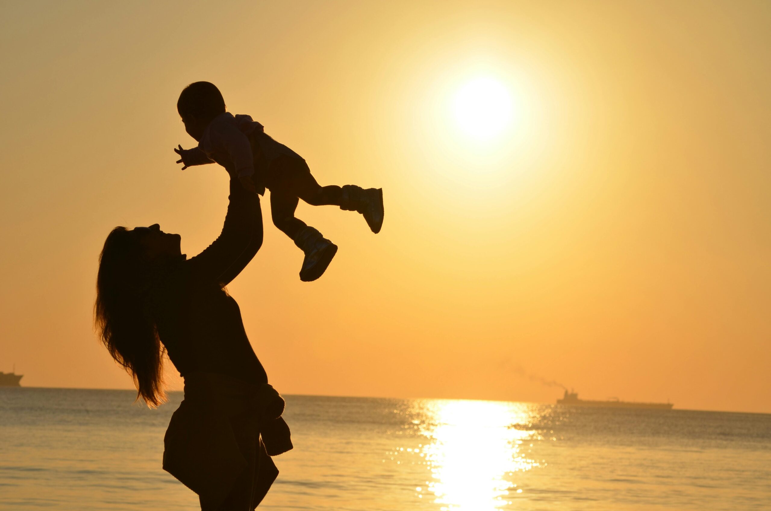 Silhouette of an adult lifting a child into the air at De Bakke Santos beach during sunset, with the sun low over the water and ships in the distance—a perfect moment for a Mother’s Day gift or unforgettable coastal escape. @De Bakke Santos Mossel Bay
